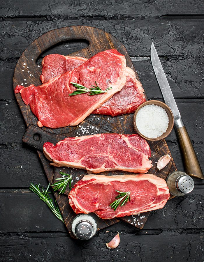 Frische, rohe Steaks auf einem rustikalen Holzbrett, dekoriert mit Rosmarin, Meersalz und Knoblauch